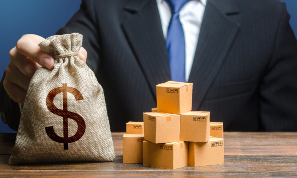 Businessman holds dollar money bag and boxes with goods. Purchase and sale of goods, trade and commerce. Import and export. Business income. Payment of taxes and fees. Advertising budget